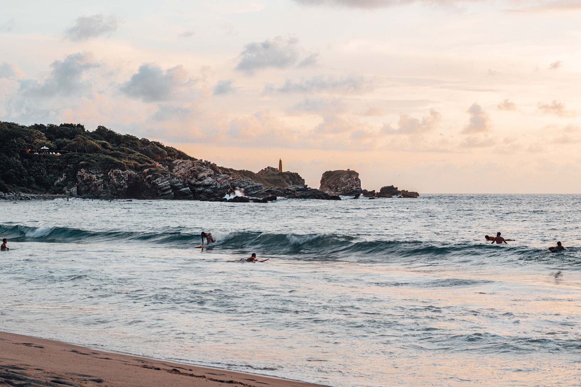Surfers in the waves off a rocky coast at sunset